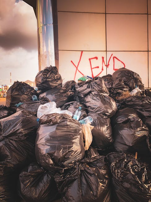 A pile of mixed rubbish including flattened cardboard boxes, brown paper bags, and plastic packaging, placed on the ground against a brick wall and the base of a tree with visible bark texture. The pa