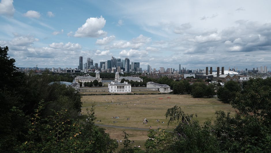 A wide panoramic view of Greenwich Park in London shows a large historic white building with classical architectural features, including multiple towers with domed roofs and an extensive colonnaded te