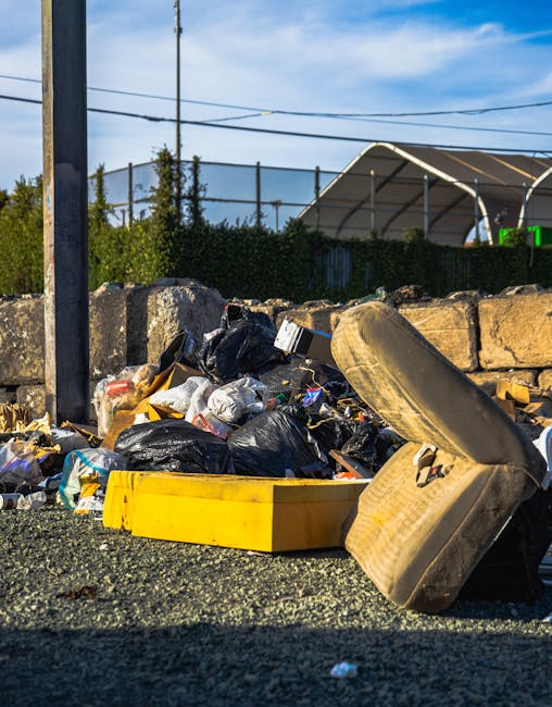 A patch of ground with scattered rubbish including torn cardboard boxes, some with green and white branding, and crumpled paper. The debris is spread across a grassy area near the edge of a fence, wit